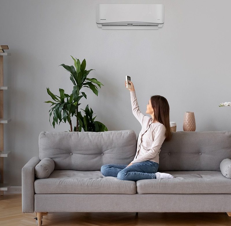 Woman on a sofa adjusting a wall-mounted heat pump with a handheld remote