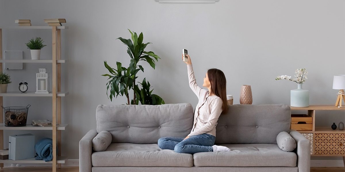 Woman on a sofa adjusting a wall-mounted heat pump with a handheld remote